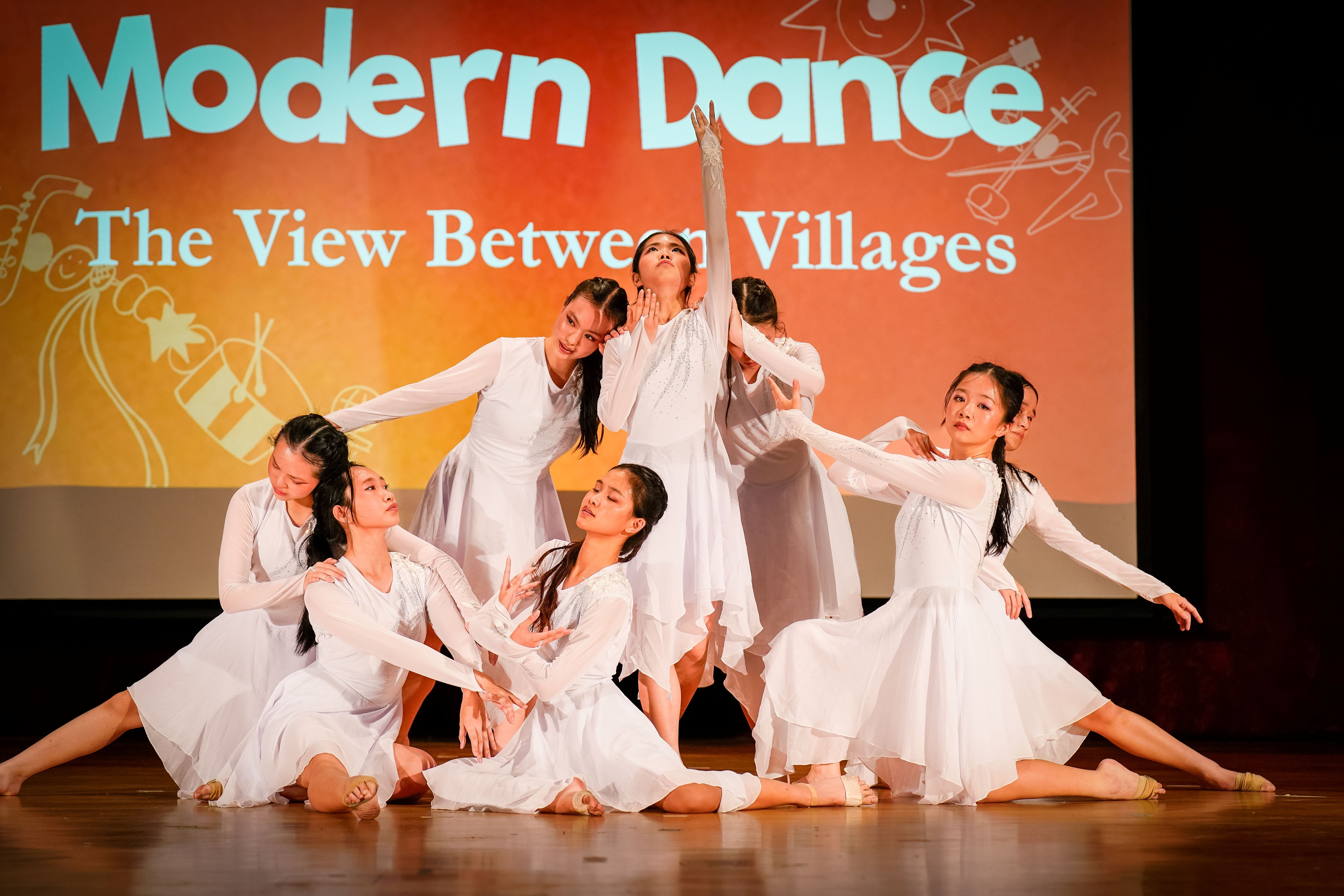 A group of dancers in white dresses perform a graceful formation on stage, with one dancer reaching upward as others pose around her.